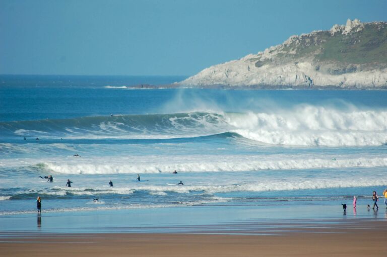A family walks on a dog friendly beach in Devon whilst people surf in the water
