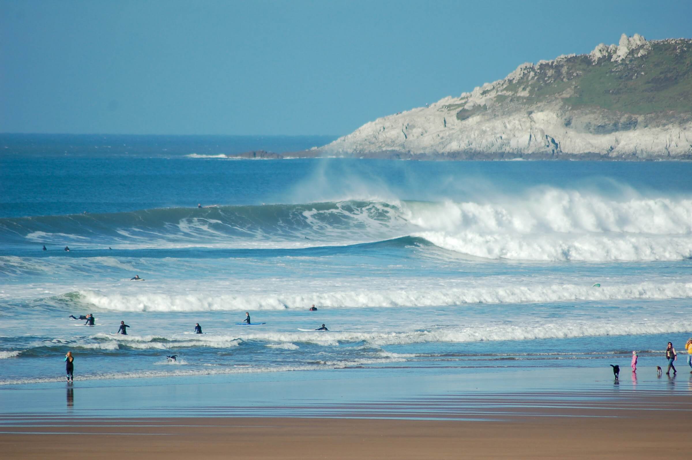 A family walks on a dog friendly beach in Devon whilst people surf in the water