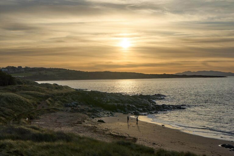 A couple walk their dog in the surf on a dog friendly beach in Scotland during sunset